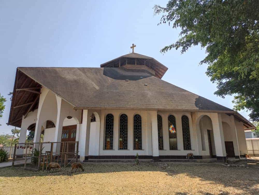 A white chapel with a dark roof on a field with goats grazing out front.